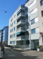 Modern apartment building with multiple balconies, white exterior, blue awnings, and a fenced entrance.