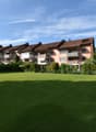 A row of apartment buildings with brown roofs and balconies, set against a backdrop of greenery and a wide grassy field.