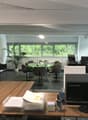 The image shows a modern and spacious office space with large windows providing a view of the surrounding greenery. The office features a glass-enclosed meeting area with a round table and comfortable chairs. There is also a desk with a Lenovo computer an