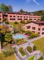 Multi-story brick building with red tile roofs, surrounded by lush gardens, landscaping, and a swimming pool. The building has balconies and terraces overlooking the grounds.