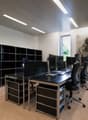 The image shows a modern office space with multiple workstations. The room has a sleek and minimalist design, with black desks, chairs, and storage units. There are several computer monitors and printers on the desks, indicating this is a workspace for of