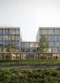 Modern office building with glass facade, multiple floors, entrance with glass doors, people walking around, trees, and green lawn