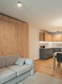 Open-plan living and dining area with a gray sofa, dining table, and wooden flooring. The kitchen is partially visible with a sink and cabinets.