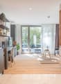 The image shows an open concept living space with a kitchen, dining area, and living room. The kitchen features modern gray cabinets, wooden shelves, and stainless steel appliances. The dining area has a small table and chairs, and the living room has a c