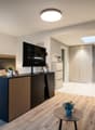 Spacious bedroom with a large bed, wooden furniture, a TV stand, and a seating area. The room has hardwood flooring and a skylight. There is also a small table and a dishwasher visible in the room.