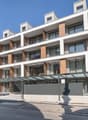 Multi-story apartment building with modern, minimalist design. The building features balconies on each floor, with a mix of glass and wood accents. There is a covered entrance area at the ground level.