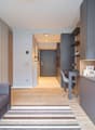 Open-plan studio apartment with living room, kitchenette, and dining area. Gray and wood tones throughout, with a gray sofa, wooden floors, and built-in shelving and storage. Entrance hallway leads to the apartment. Dishwasher visible in the kitchenette a