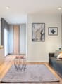 Spacious living room with hardwood floors, white walls, large windows, gray sofa, TV stand, and decorative artwork on the walls. The room also features a sliding door leading to a balcony or terrace.