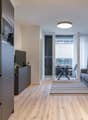 Open-concept living area with kitchen, living room, and balcony. Modern kitchen with built-in appliances including a dishwasher. Hardwood flooring throughout. Cozy living room with sofa and dining table. Large windows providing natural light and access to