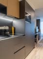Modern kitchen with wood-tone cabinets, stainless steel appliances including a dishwasher, and a potted plant on the counter. The kitchen opens up to a dining area with a glass table and chairs.