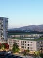Two high-rise buildings, parking area, green grass, street with tram and cars, trees, green roof on one building, mountains in the distance.