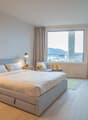 Spacious bedroom with large windows offering a scenic view, hardwood flooring, a king-size bed with storage drawers, two bedside tables with lamps, a desk with a chair, and a flat-screen TV mounted on the wall.