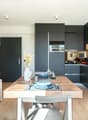 The image shows an open-plan living room and kitchen area with a large wooden dining table, black kitchen cabinets and appliances, and a cozy living space with shelving and decor. The kitchen appears to have a dishwasher visible as one of the appliances.