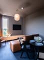 Spacious living room with large windows, brown curtains, wooden TV stand, leather sofa, dining table with chairs, and decorative elements like a fan-shaped wall art piece.