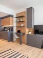 The image shows a modern and compact studio apartment with an open-plan layout. The kitchen area features dark gray cabinets and a wooden countertop, with a dishwasher visible. There is a desk area with shelves above it, and a TV stand with storage units.