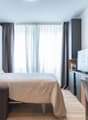 Large bed, decorative pillows, white blanket, wooden walls, white curtains, desk, monitor, cabinet, chair, shelves with frames, flower arrangement, and mug