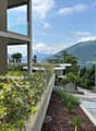 Modern building with large windows overlooking a scenic lake and mountains. The building has a balcony or terrace area with lush greenery and landscaping. The surrounding area features a paved walkway and additional landscaping elements.