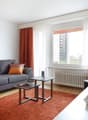 Spacious living room with a large gray sofa, orange accent pillows, and a framed artwork on the wall. The room has a large window with red curtains, providing a view of the city outside. There is a coffee table and a TV stand with a flat-screen TV.