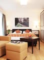 Spacious bedroom with hardwood flooring, large windows, a king-size bed with striped bedding, a seating area with two yellow armchairs and ottomans, a TV stand, and a framed artwork on the wall.