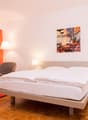 Spacious bedroom with a large bed, wooden floor, orange curtains, side tables with lamps, and a framed artwork on the wall depicting a city scene.