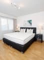 spacious bedroom with large bed, lamps, side tables, nightstands, white linens, framed photo, wooden floor, large window with white curtains