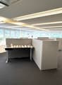 The image shows a modern office space with a large open floor plan. The space has a reception desk, cubicles, and various storage units. The room has large windows that provide natural lighting, and the ceiling has exposed beams and lighting fixtures. The
