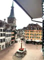 Church clock tower, street with shops, square with fountain, buildings, cobblestone streets, pedestrians
