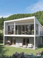 Two-story modern house with a flat roof, large windows, and a balcony on the upper level. The house is surrounded by a well-manicured lawn and lush greenery, including trees and flowering plants. There appears to be a garage attached to the side of the ho