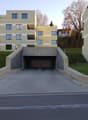 Multi-story apartment building with a garage entrance in the foreground. The building has a modern, minimalist design with a mix of yellow and gray colors. There are balconies and large windows visible on the facade. The surrounding area appears to be a r