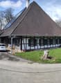 A traditional German-style half-timbered house with a large pyramidal roof, surrounded by trees and a grassy area. There is a parked car in the driveway in front of the building.