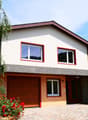 2-story house with white exterior walls, red-framed windows, and a wooden garage door. The house has a balcony or terrace on the upper level. The front yard features a paved driveway and red flowers in the landscaping.