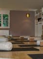 A yoga studio with yoga mats, bolsters, pillows, and lamps on the ceiling. The floor is wooden, and the walls are painted in a muted purple. There is a shelf with towels, a potted plant, and a framed artwork.