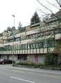 Multi-story apartment building with balconies, brick and concrete construction, surrounded by trees and vegetation