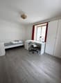 room features a single bed, desk with a chair, a window with red curtains, and a lamp. White walls and wooden floors.