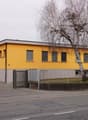 A single story yellow building with gray trims. A gate leads to the parking lot with trees in the center. Several windows and a chimney are visible on the yellow wall. A grey building is on the right side.