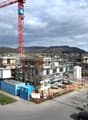 Aerial view of a construction site with multiple buildings under construction, a large red crane, scaffolding, and a road with parked cars.