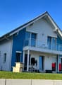 Two-story modern house with white exterior and blue accents, large windows, balcony, and attached garage.