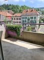 Empty balcony with concrete floor and railing, overlooking a street with trees, houses and hills.