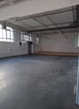 Large empty warehouse room, white walls, metal flooring, fluorescent ceiling lights, windows, radiators, small raised platform, door