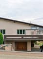 2-story house with stucco exterior, brown roof, large windows, wooden garage door, balcony, and a parked car in the driveway. The house is surrounded by landscaping with shrubs and trees.