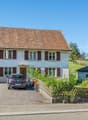 Two-story house, brown roof, large windows, balcony, garage, two cars parked in driveway