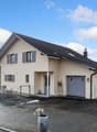 Modern house, beige exterior, black roof, large windows, two garages, driveway, fenced garden, mountains in the distance