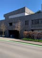 Modern building, gray exterior, multiple windows, entrance with garage, trees, bushes, road