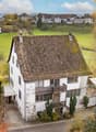 A white house with a balcony, brown roof, situated in a village, surrounded by green fields and mountains.