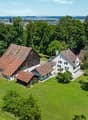 Two-story farmhouse with a red tile roof, surrounded by lush green trees and a large grassy lawn. The property also includes a smaller wooden barn-like structure. The houses have white walls and windows, and there appears to be a balcony or terrace on one