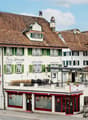 A multi-story building with a traditional European architectural style, featuring red-tiled roofs, green shuttered windows, and a balcony. The building houses a tea room on the ground floor, with outdoor seating. The surrounding buildings have a similar s
