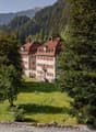 Large historic manor house with red-tiled roofs surrounded by lush green forests and mountains in the background. The house appears to be situated on a grassy field with a stone wall in the foreground.