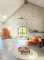 Spacious open-concept living area with high ceilings, arched windows, concrete walls, and a fireplace. The room features a mix of rustic and modern decor elements, including a large orange sofa, wicker furniture, and decorative accents like a hanging plan