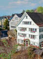 Aerial view of a row of houses with brown roofs and white walls, surrounded by trees and plants, with a tower in the distance.
