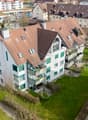 Aerial view of a multi-story residential building with brown roofs, white walls, and green window frames. Balconies are visible on the upper floors. The building is surrounded by a well-maintained green lawn and several trees. A car is parked in the parki